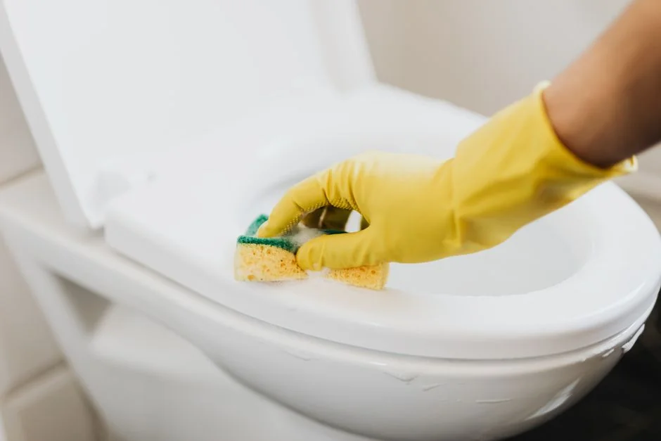 An unidentified individual wearing bright yellow rubber gloves scrubs a bathroom mirror with a basic sponge